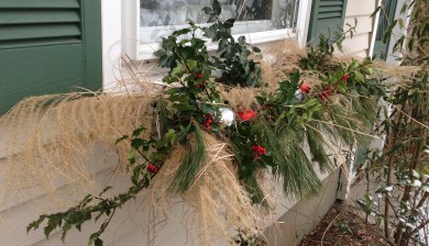 A winter window box decorated with holly with red berries, white pine branches, dry ornamental grasses, and red and white Christmas balls.