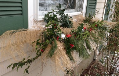 A winter window box decorated with holly with red berries, white pine branches, dry ornamental grasses, and red and white Christmas balls.