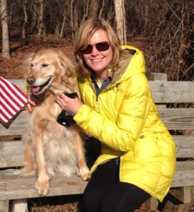 Mary Stone wearing a yellow jacket sitting on a bench with Ellie her gold retriever at Henricus Historical Park
