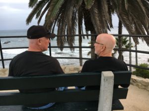 two men in black shirts and bald heads sitting on a bench overlooking he ocean