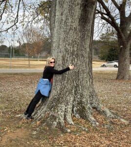 a blonde woman, Mary Stone, in sunglasses and a denim shirt hugging a three foot wide water oak trunk.