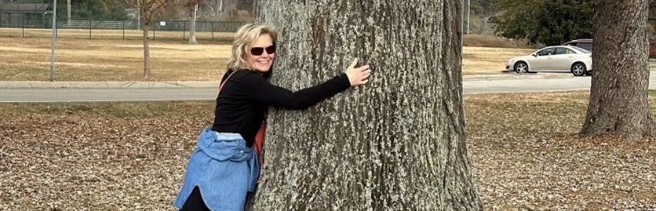 a blonde woman, Mary Stone, in sunglasses and a denim shirt hugging a three foot wide water oak trunk.