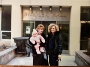 Two blind women, one holding a baby, in front of building labeled Naglar Hall. 