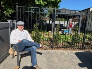 a man in a white shirt, cap and jeans sitting in front of a fence and garden tweaks underway.