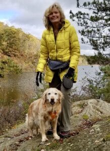 Blonde woman in a yellow coat wiht a golden retriever on a leash out in the woods