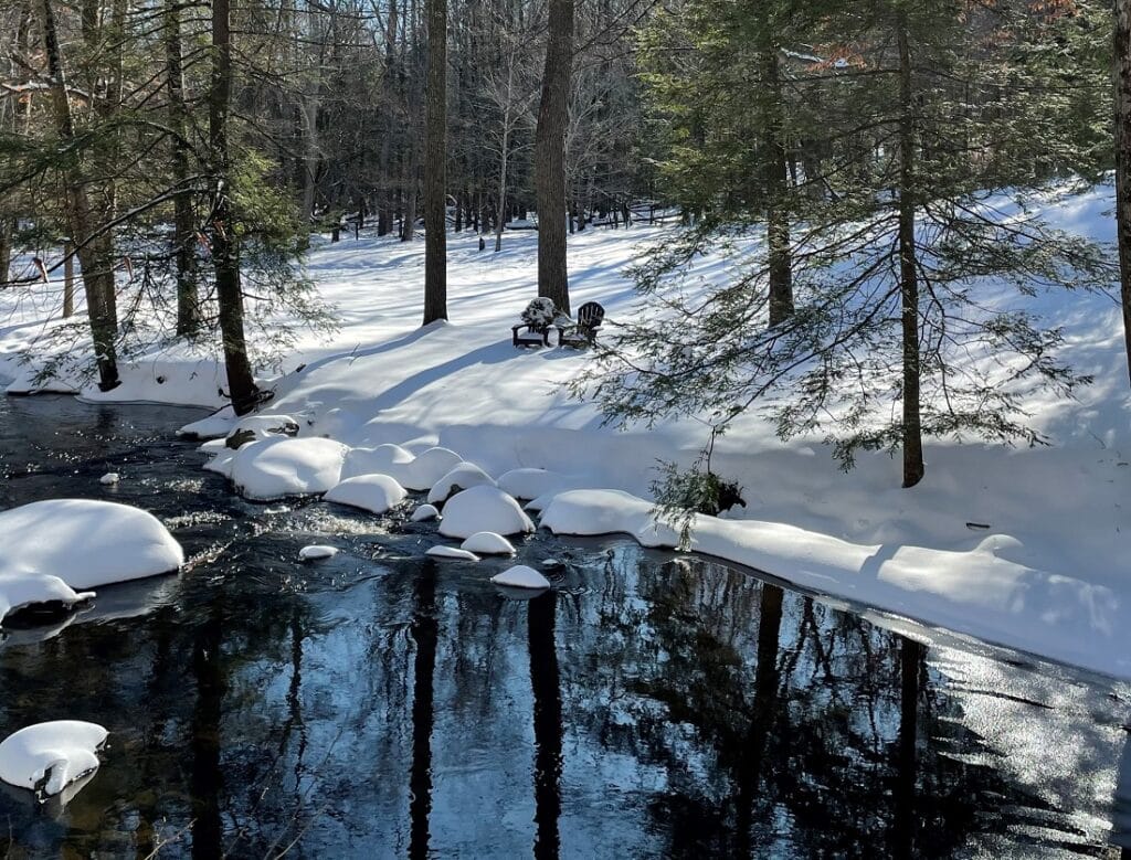 two brown chairs in snow in front of a brook with reflections of the trees