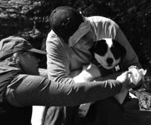 a balck and white photo of a man in a ballcap holding a black and white dog and a woman wiping the dogs paw.