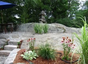 a massive grey outcropping of boulders with an Inuksuk as a stacked stone bridge over a water feature with purple echinacea, lamb's ear and fountain grass in front
