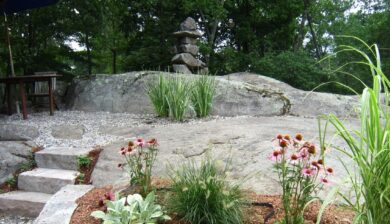 a massive grey outcropping of boulders with an Inuksuk as a stacked stone bridge over a water feature with purple echinacea, lamb's ear and fountain grass in front