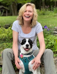 a blond woman in a grey t-shirt sitting with a black and white dog both smiling