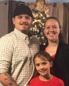 a mustached man wearing a black cap next to his brown haired wife and daughter in front of Christmas tree.