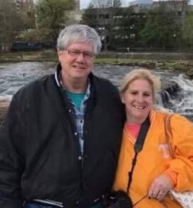 a tall grey haired man in glasses wiht a blond woman in an orange jacket holding a camera.