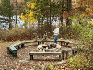 a woman with two dogs sitting on a circle of wooden benches in front of a fire pit