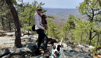 Mary Stone on the outcropping at Blue Mountain Lake carrying Callie, a small brown dog, with Jolee, a large white and back dog at her feet.