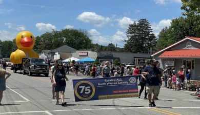 a man and woman carrying a blue banner of the Blairstown Rotary followed by a yellow rubber duck float at the town Memorial Day Parade