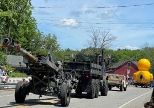 a yellow rubber duck float leading a historic military vehicle towing an enormous cannon.