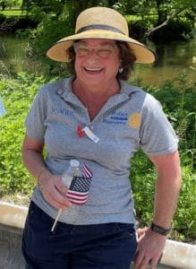 A woman wearing a straw hat and grey shirt wihth the Blairstown Rotary logo carrying a small American flag