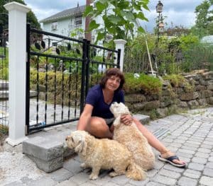 Rosemary DeTrolio, wiht dark shoulder length hair and a blue shirt, sitting on a garden step with her two tan dogs