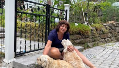 Rosemary DeTrolio, wiht dark shoulder length hair and a blue shirt, sitting on a garden step with her two tan dogs