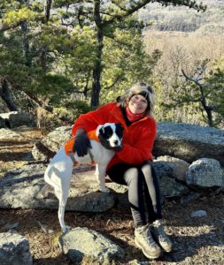 Mary Stone with Jolee, a white dog with black markings, sitting on a rock at a viewpoint at Blue Mountain Lake