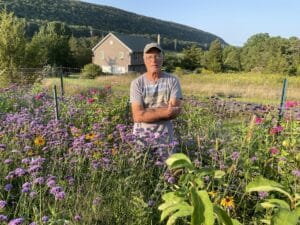 Dennis Briede in a ball cap standing in his meadow of colorful plants with a mountain ridge behind him.