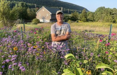 Dennis Briede in a ball cap standing in his meadow of colorful plants with a mountain ridge behind him.
