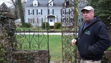 Bruce Crawford in a ball cap and black fleece in front of an iron gate and the main house at Willowwood Arboretum