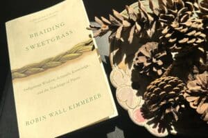 A book, Braiding Sweetgrass, with a pale yellow over and braded sweetgrass next to a plate of pinecones.
