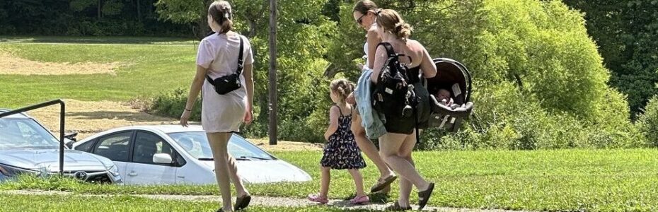 a little girl in a blue sundress and pigtails walking with a group of ladies.