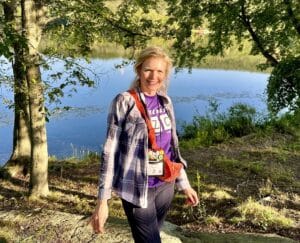 Mary Stone in front of a lake wearing a purple Comfort Zone Camp T shirt.