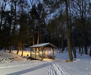 a covered footbridge in an early morning snowy scene with foot steps, a slivered moon peaking through hemlock trees,