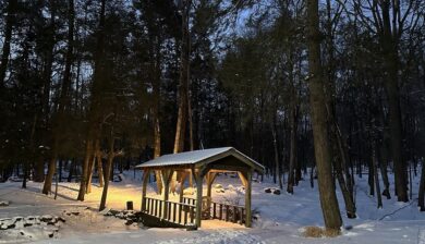 a covered footbridge in an early morning snowy scene with foot steps, a slivered moon peaking through hemlock trees,