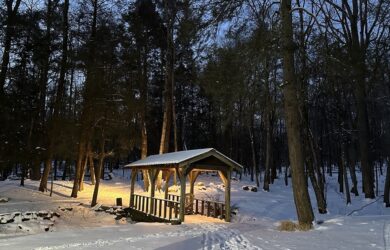 a covered footbridge in an early morning snowy scene with foot steps, a slivered moon peaking through hemlock trees,