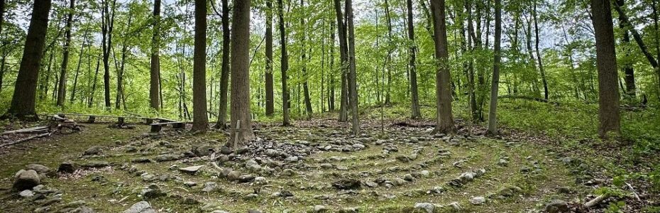 a stone edge maze amongst a forest with an upright wooden cross in next to a tree in the center.