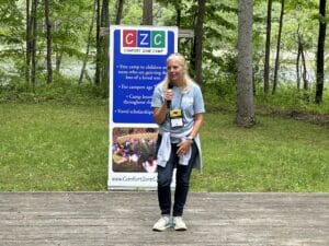 Lynne Hughes, found of Comfort Zone Camp, with blond hair and jeans holding a microphone in front of a CZC banner.