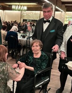 John Quinlan wearing a tuxedo behind Julia Quinlan in a green sparkly dress sitting in a wheel chair.