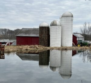 Red barns and tall silos reflected in calm winter pond water create a peaceful rural landscape of symmetry, stillness, and quiet seasonal beauty.