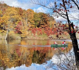 Two paddlers in a green canoe glide across a reflective autumn pond surrounded by brilliant fall foliage and shimmering mirrored color.