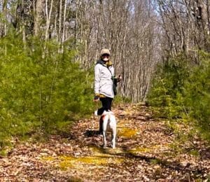 Mary Stone and her rescue dog Jolee standing on a woodland trail surrounded by young evergreen trees.