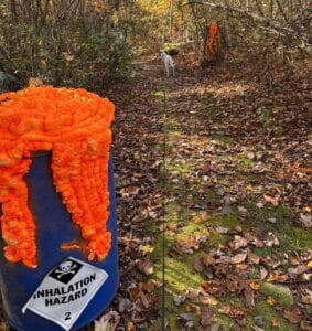 Blue barrel with an inhalation hazard warning and bright orange Halloween decorations beside a wooded trail.