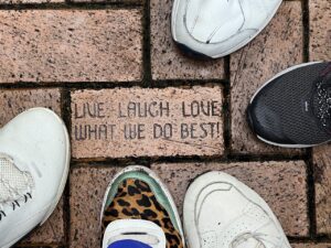 Engraved brick reading “Live Laugh Love What We Do Best!” surrounded by visitors' shoes.